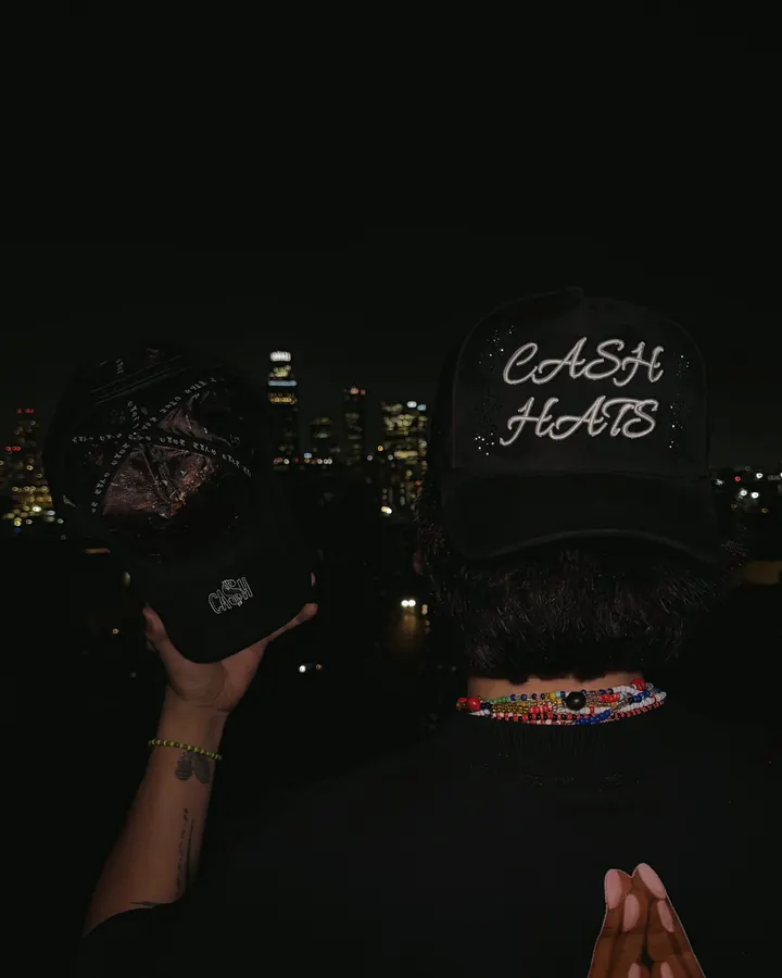 Una mano sostiene la gorra negra CASH HATS con el skyline de Los Ángeles de noche al fondo