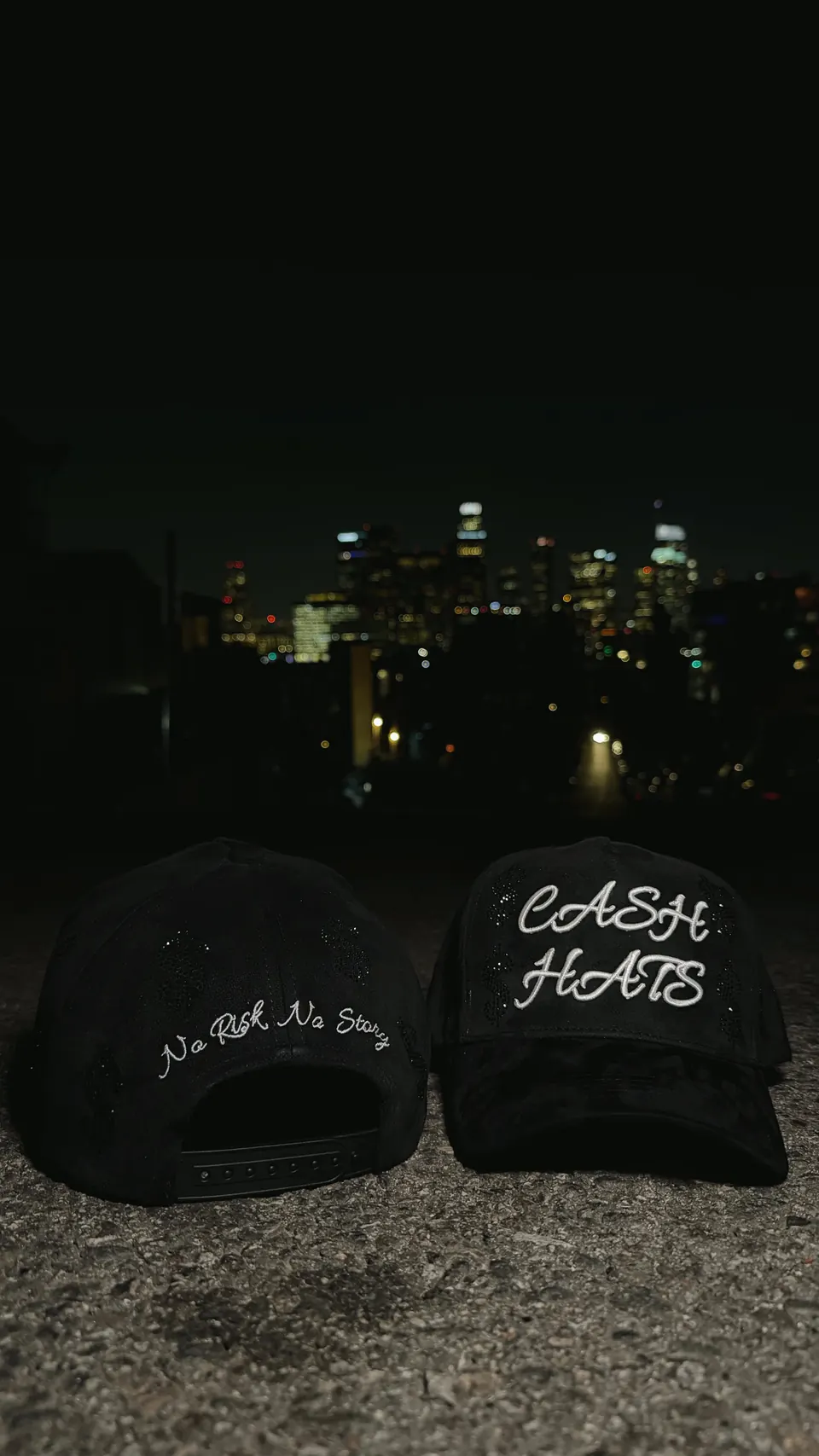 Dos gorras negras CASH HATS en el asfalto frente al skyline de Los Ángeles: una con “CASH HATS” bordado al frente y otra con “No Risk No Story” atrás