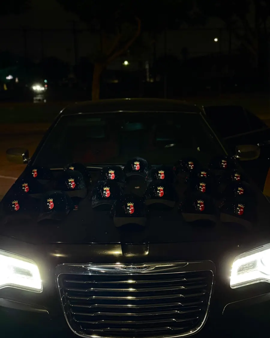 Gorras negras DROP ELEGUA con el parche de Elegua rojo y negro, acomodadas sobre el cofre de un carro con los faros encendidos