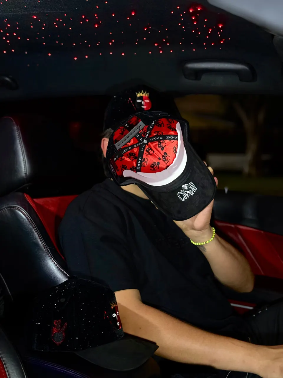 Un hombre en un carro muestra el interior rojo estampado de la gorra DROP ELEGUA y el bordado CA$H bajo la visera