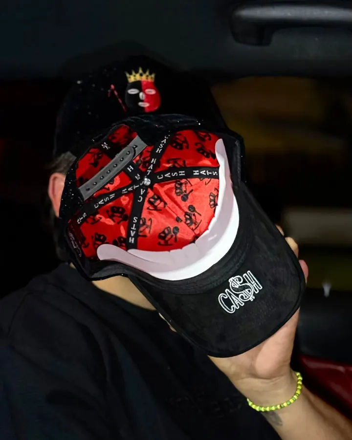 Un hombre en un carro muestra el interior rojo estampado de la gorra DROP ELEGUA y el bordado CA$H bajo la visera