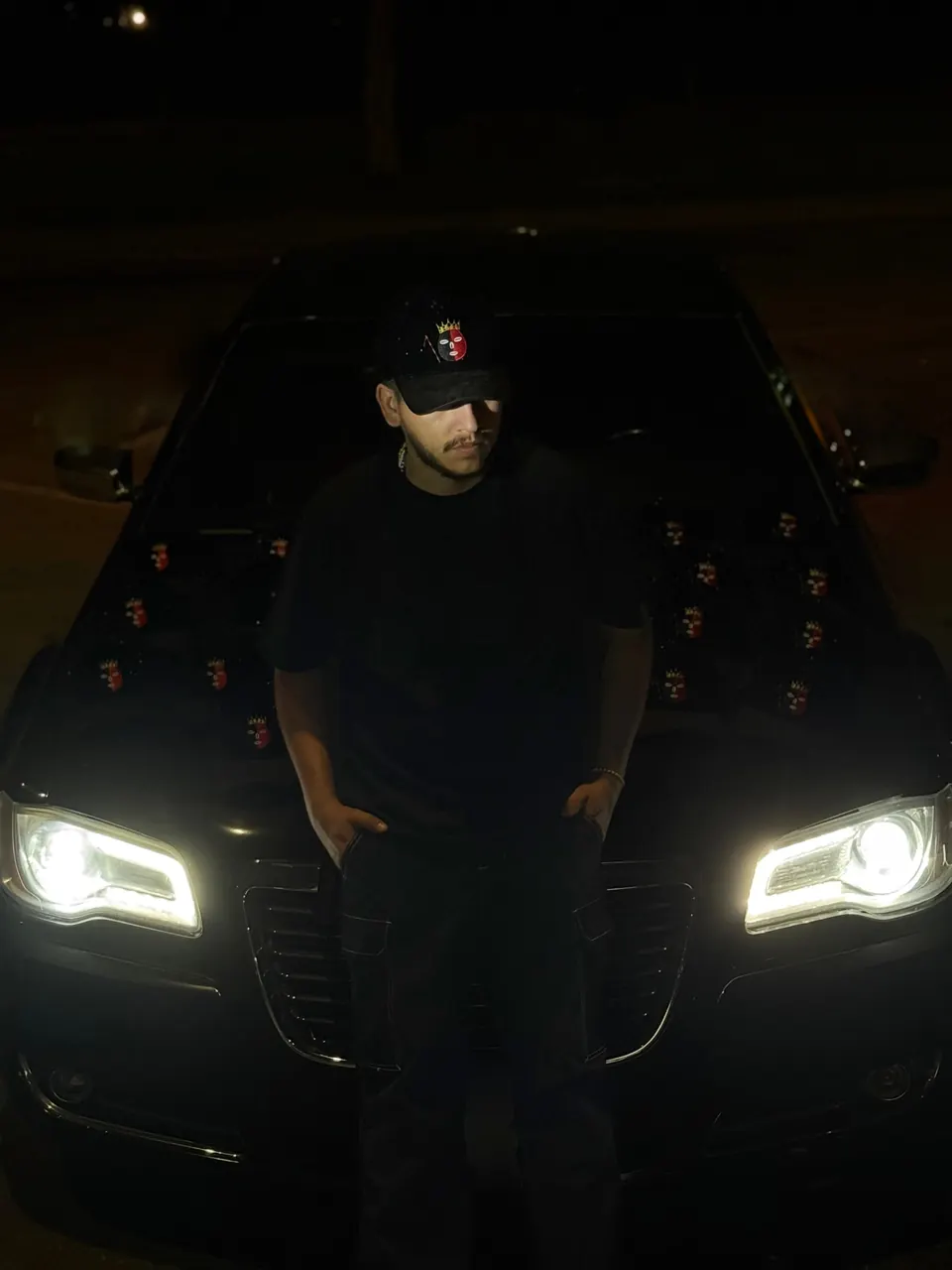 Un hombre de pie frente a un Chrysler negro con las luces encendidas y varias gorras DROP ELEGUA sobre el cofre