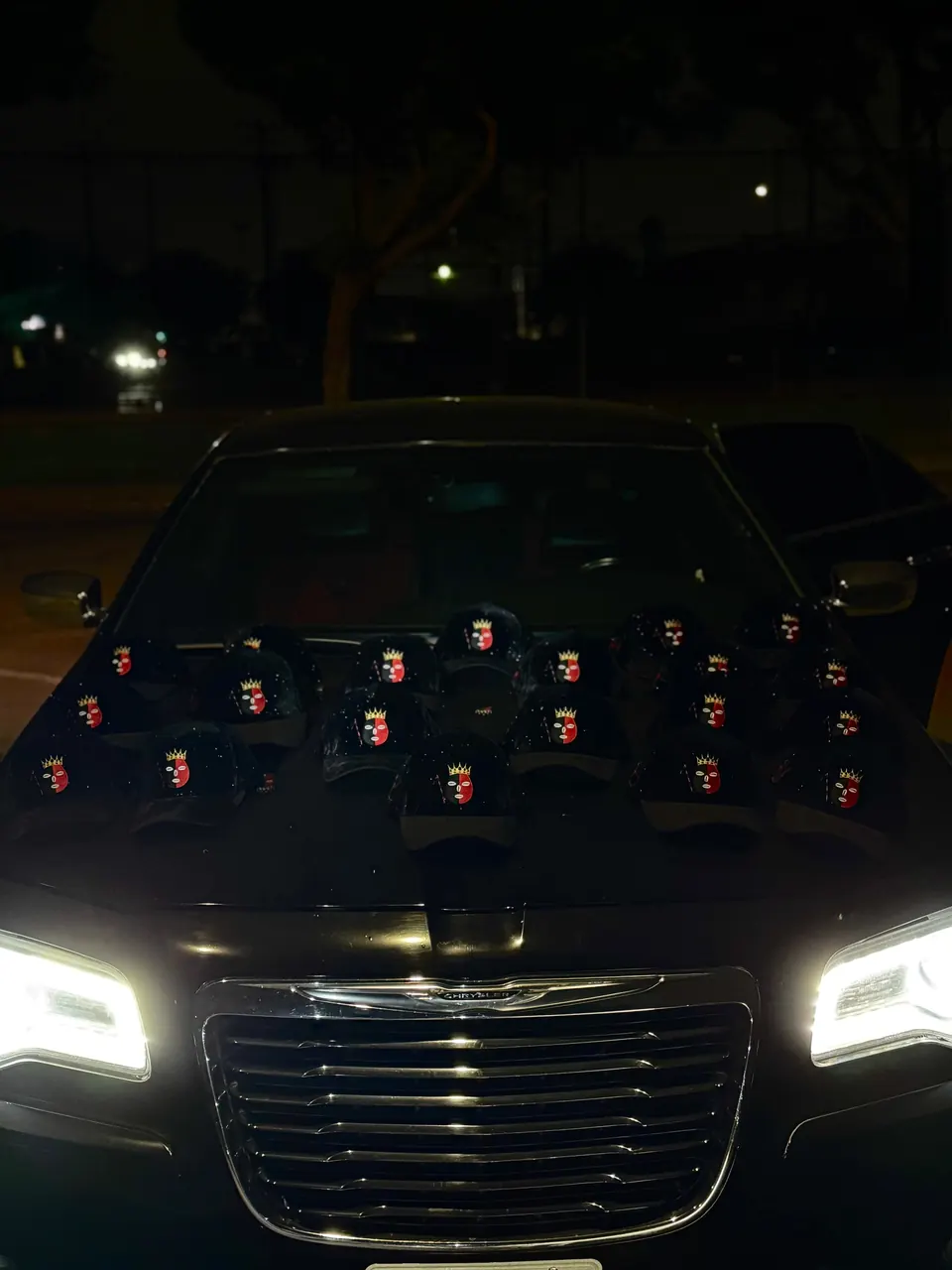 Gorras negras DROP ELEGUA con el parche de Elegua rojo y negro, acomodadas sobre el cofre de un carro con los faros encendidos