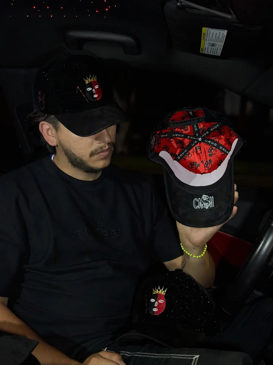 Un hombre en un carro lleva la gorra DROP ELEGUA con el parche de Elegua y la corona dorada, y sostiene otra mostrando el forro rojo