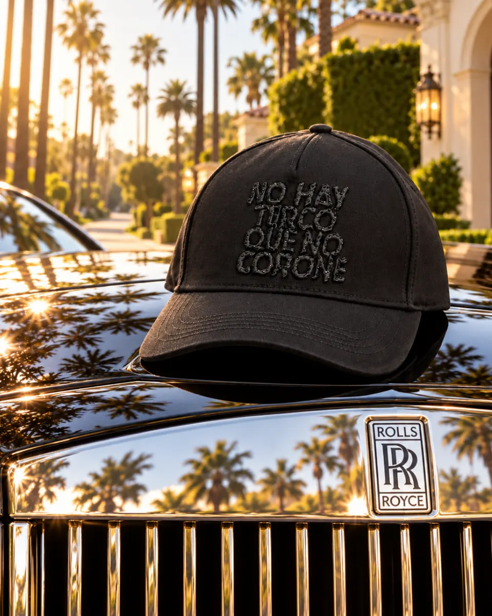 La gorra NO HAY TERCO QUE NO CORONE sobre el cofre de un carro negro, con palmeras y luz dorada al fondo